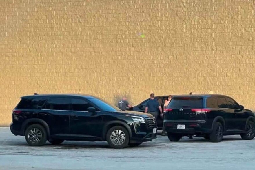  Three black unmarked SUVs parked next to a blank exterior wall with several people standing outside one of them closest to the wall. 