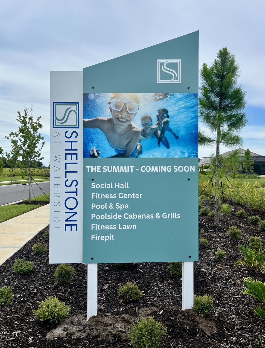 A sign outside the Shellstone at Waterside community. It reads: the summit coming soon. Social hall, fitness center, pool and spa, poolside cabanas and grill, fitness lawn and firepit. It shows a group of kids swimming under the water. 