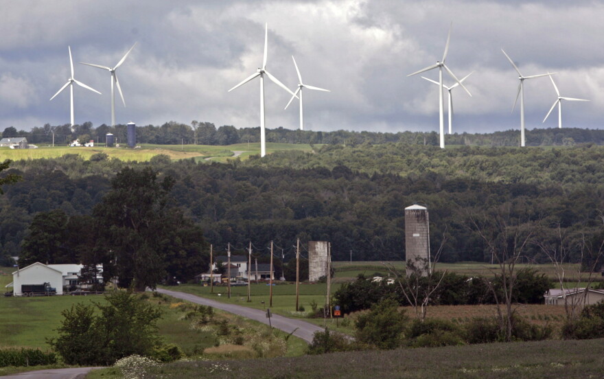 A rural landscape. Farms and trees are visible in the foreground. In the background are 8 large white windmills.