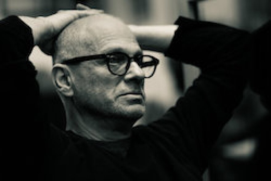 Black and white image of composer David Lang in glasses gazing away from the camera in thought, with his hands crossed behind his head