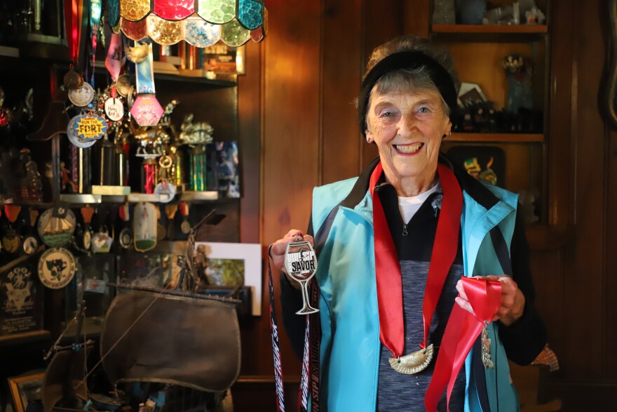 An elderly woman wearing workout gear and a medal is smiling and holding two medals. Behind her is a trophy case filled with awards.