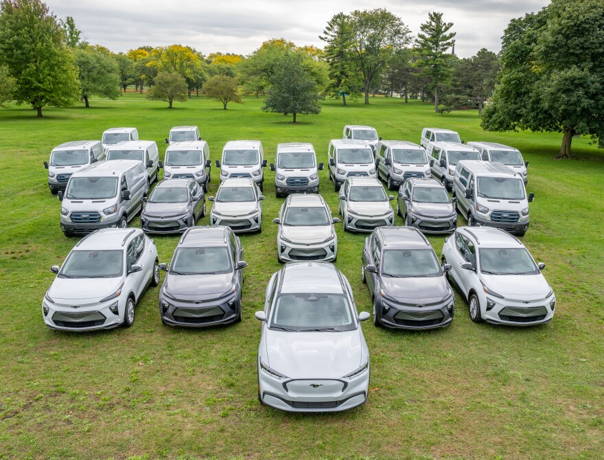 25 electric gray and white cargo vans and cars parked side by side in several rows on a green lawn.