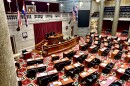 A view from the gallery of the Missouri House of Representatives floor. Empty wooden desks and dark chairs face the dais. 