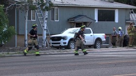 Firefighters walking away from a house with two ladders leaning on it. Three other men in firefighting gear are talking near the house.