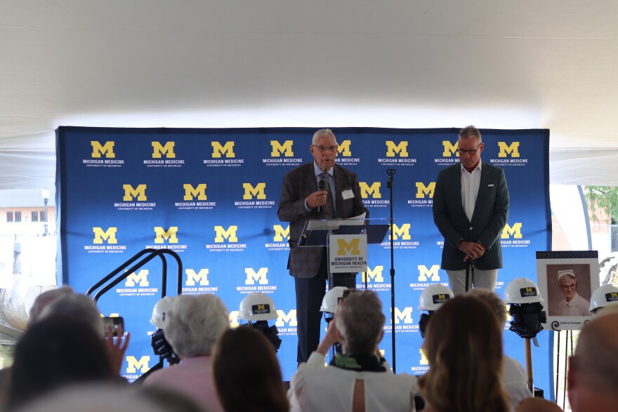 An older man stands at a podium on stage, addressing the audience. A man in a gray suit stands next to him.