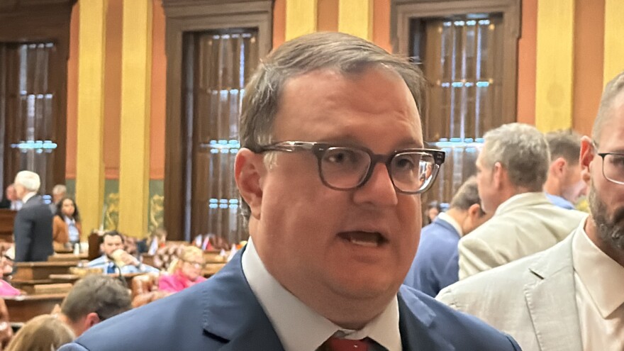  A man wearing glasses, a blue suit jacket, white dress shirt, and red tie speaks in a legislative chamber. He is photographed in close-up from a slightly lower angle, mouth open mid-speech. Behind him, the ornate interior of the Michigan State Capitol is visible — warm yellow and terra cotta walls, dark wood paneling, and a crowded floor with other people in business attire milling about.