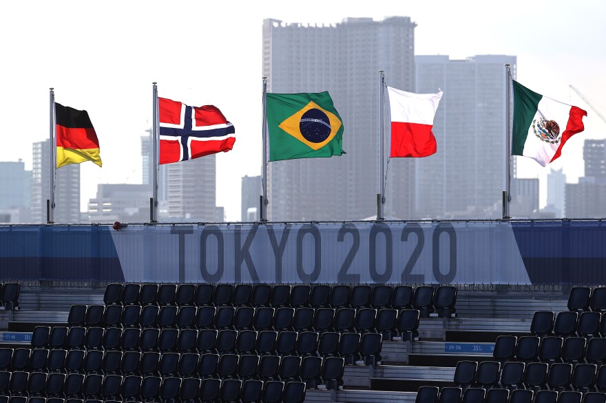 A sign reading "Tokyo 2020" and five international flags are visible above an empty set of bleachers, in front of a skyline of grey high-rise buildings.