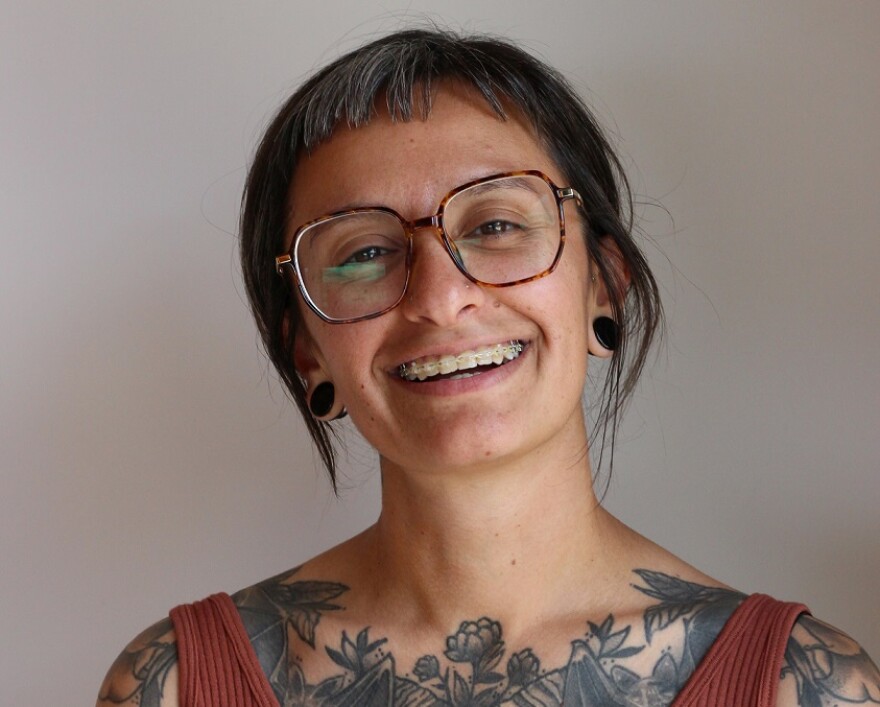 Dima Khalifa smiles. She has floral ttatoos across her chest, grazing her collarbone, and shoulders and wears an orange ribbed v-neck tank top.  