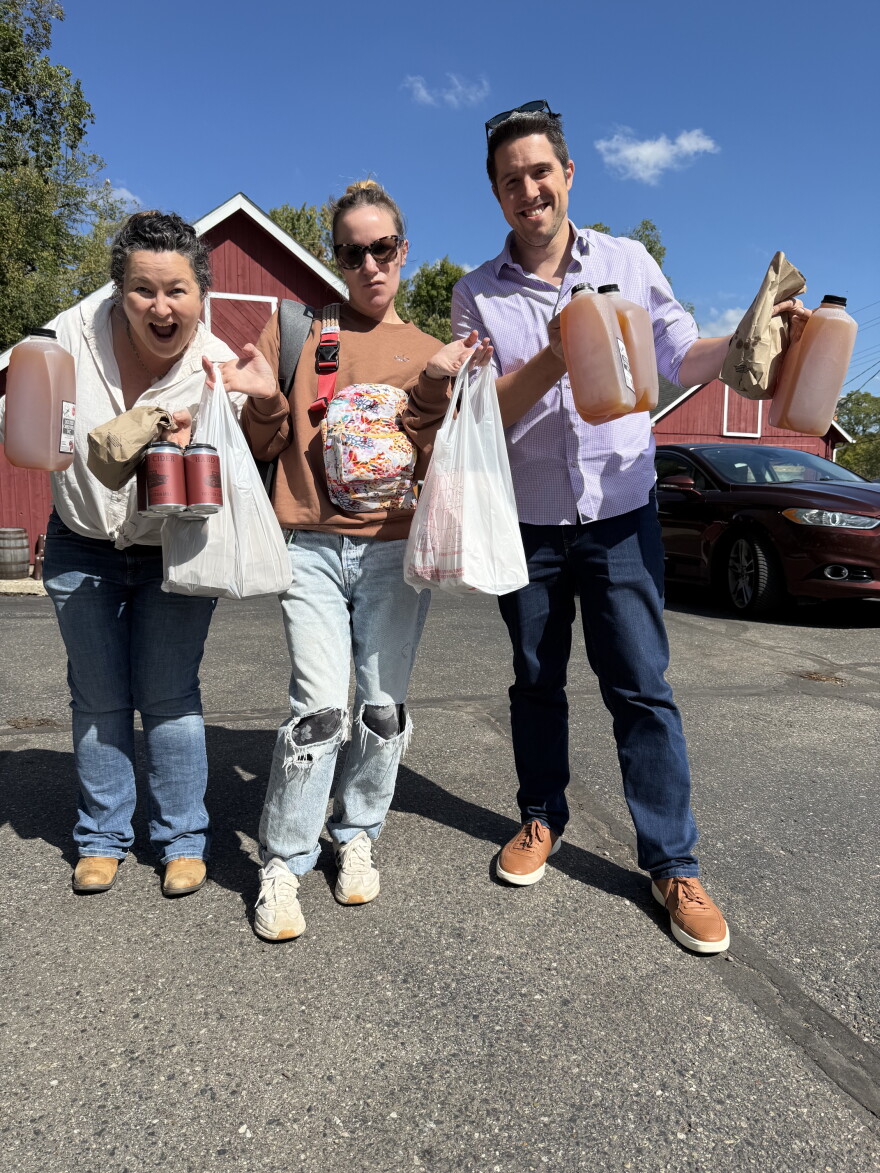 three people stand holding jugs of cider and bags of doughnuts in front of a red barn