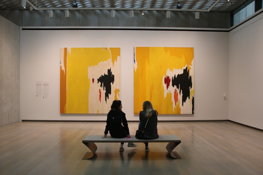 Two women sit on a bench. In front of them are two similar paintings next to each other, primarily yellow with black splotches of paint on the right. The painting on the left has a smaller black splotch, and the painting on the right has a larger black splotch and some red marks next to it. 
