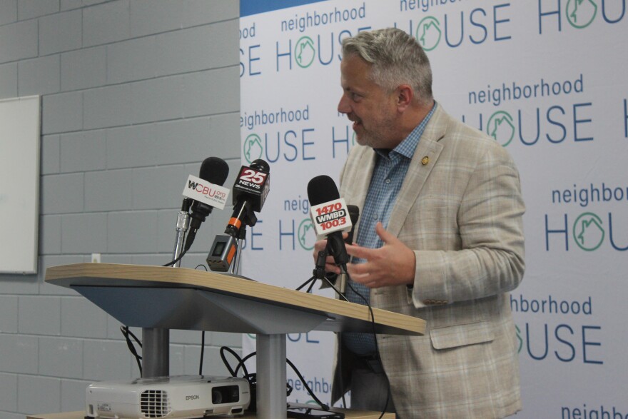 Sorensen wears a tan suit and blue shirt and stands at a podium. 