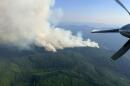 Aerial view from a fire spotting plane. A large plume of smoke rises from green, tree-covered mountains. Part of an airplane propeller is visible in the frame. 