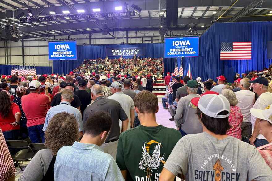 Many supports stand in a large venue with screens saying "Iowa Welcomes President Donald J Trump." 