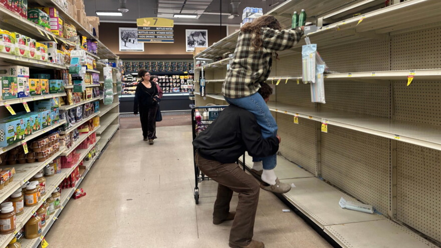A man hoists a woman on his shoulders to reach water bottles on the top shelf of an otherwise empty set of shelves.