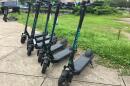 Five black e-scooters are lined on the sidewalk. The back wheel of each scooter touches the grass and the front wheel faces the road. Teal words spell "VeoRide" on the scooter's stem. Tallahassee buildings are in the distance.