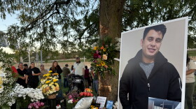 A poster-sized photo of a smiling man in a dark zip up sweater sits on an easel in front of a treet surrounded by colorful flowers. On the ground, white tea lights are arranged in the shape of a cross. 