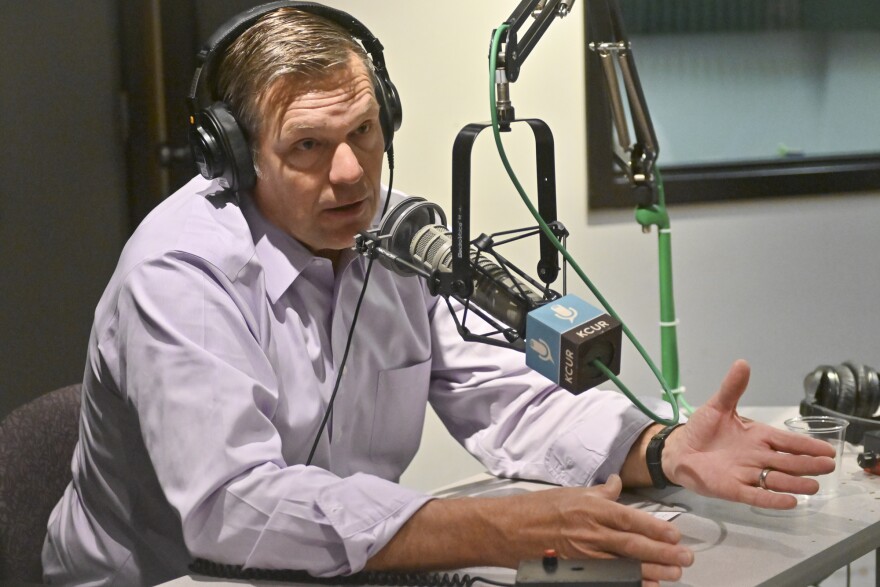 A man inside a radio studio sits behind a microphone. He is talking and gesturing with both hands.