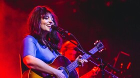 A woman smiles and sings into a microphone while playing an acoustic guitar on stage with red lighting in the background.