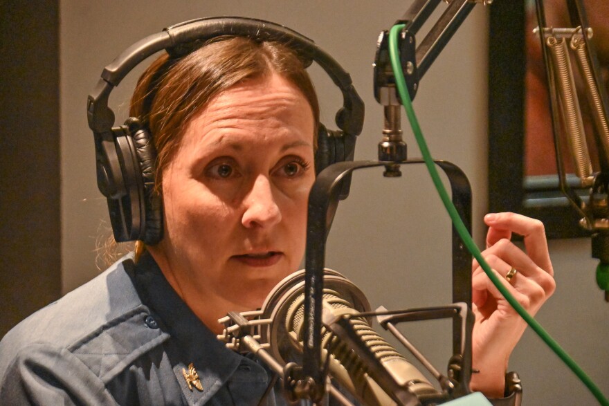 A woman wearing a police chief's uniform and headphones talks at a microphone inside a studio. She's gesturing with her left hand as she talks.