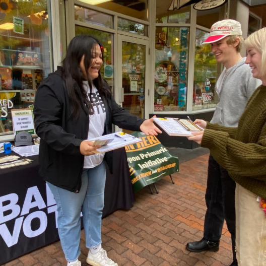 A woman wearing a t-shirt and blazer hands a clipboard to two people who are smiling. She is standing in front of a table with a black tablecloth that says "Babe Vote" 