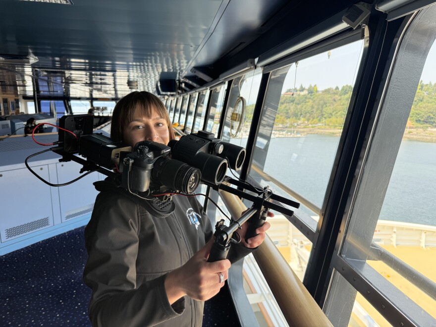A woman wearing a black jacket standing next to the picture windows on the bridge of the MSC Poesia holds a large rig that combines binoculars and a video camera. 