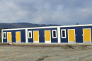 A row of small buildings with blue walls and white trim has six yellow doors and five windows. The ground in front is gravel. 