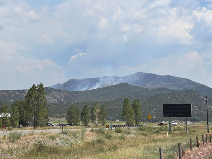 Roadside photo near brush-covered mountains. Smoke is visible behind one of the mountain's ridges.