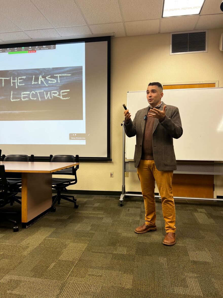 A professor in a sport coat holds a pointer as he gestures to the class while speaking. Behind him, a screen shows the words "The Last Lecture."