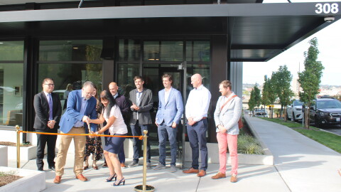 Project leaders stand in front of a new gray and white apartment complex, cutting a ribbon in front of an open entrance. 