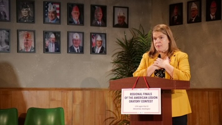 A woman wearing a yellow coat standing in front of a podium with pictures of military servicemen in the background.