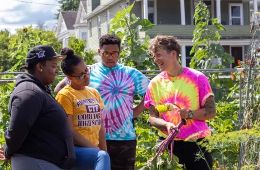 Four people stand outside conversing amidst greenery. One holds rhubarb while smiling.
