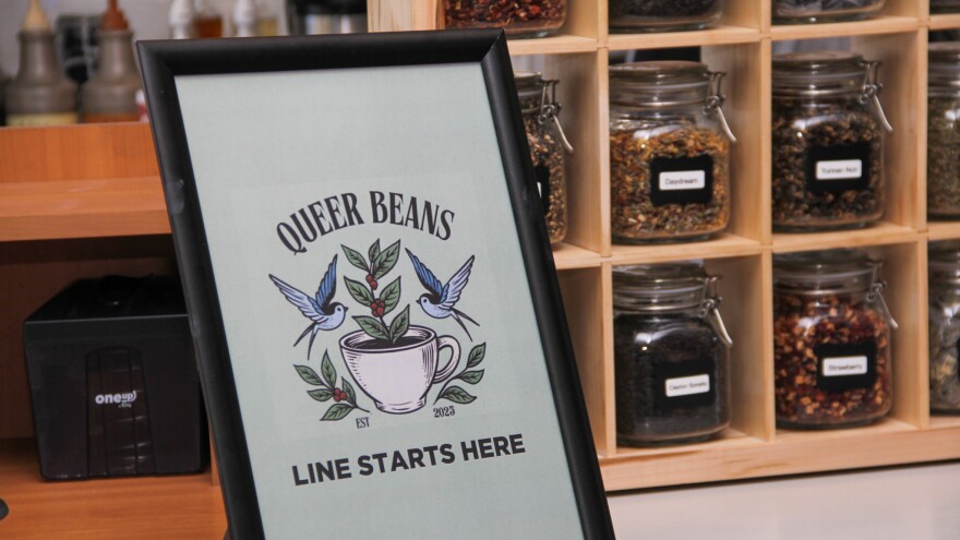A sign on the wood counter of a coffee shop reads, "Queer Beans, line starts here." The sign has a drawing of two birds flying next to a coffee mug with sprigs of leaves and berries. Behind it, small mason jars hold dry tea. 