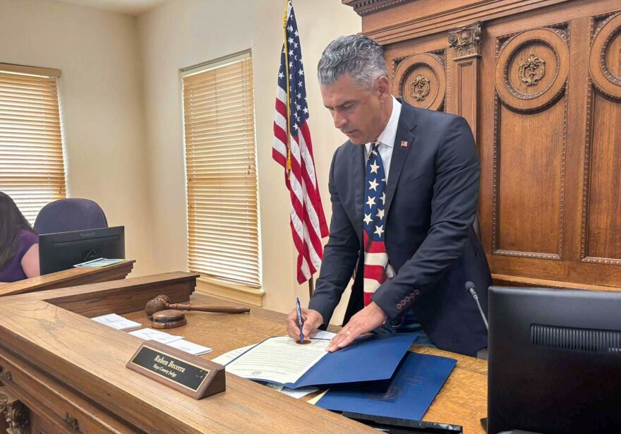 A man in a suit and American flag tie signs a paper at his seat at the head of a commissioners court meeting.