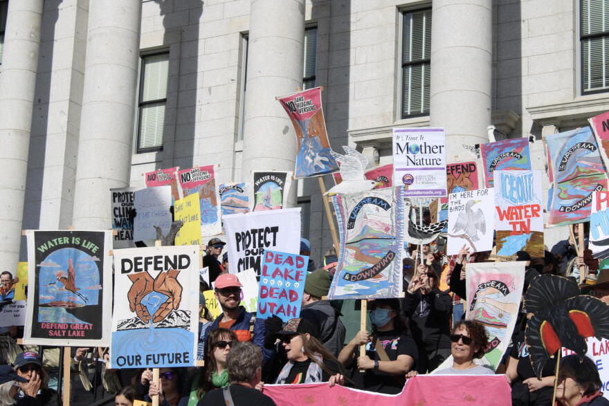 A group of people hold colorful protest signs in front of a light grey building with columns. A couple signs say "WATER IS LIFE DEFEND GREAT SALT LAKE" AND "UPSTREAM & DOWNWARD."
