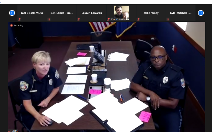Two police officers in uniform sit on either side of a long, dark table with and pink papers in piles