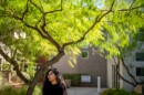 A person with long dark hair stands in front of a tree.