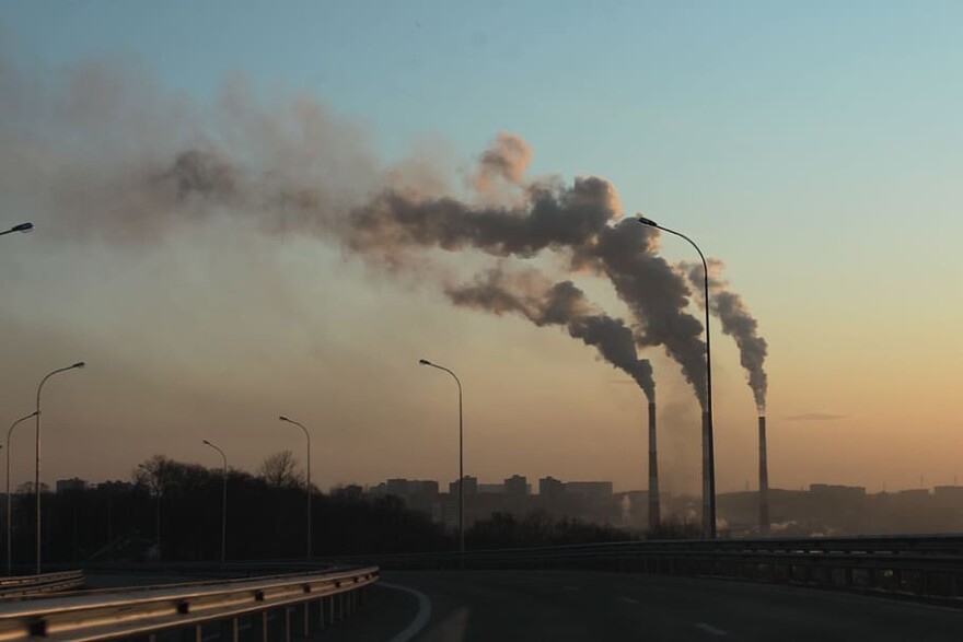 A factory with three smoke stacks is emitting smoke into the air. A road curves in front of the factory. 