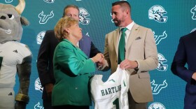 A woman in a green blazer shaking hands with a man in a beige blazer, with his other hand holding a football jersey.