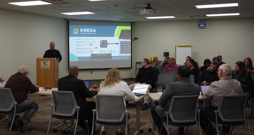 A man in a long sleeve black t-shirt stands a light brown podium, speaking to a row of trustees at the KRESA Board of Education meeting. Attendees can be seen to the right of the image, looking at the man as he talks with the board. 