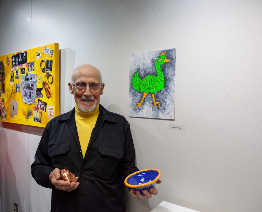 An older man with glasses stands indoors, smiling and holding pottery. Behind him, a painting of a bright green duck hangs on a white wall alongside other colorful art pieces.