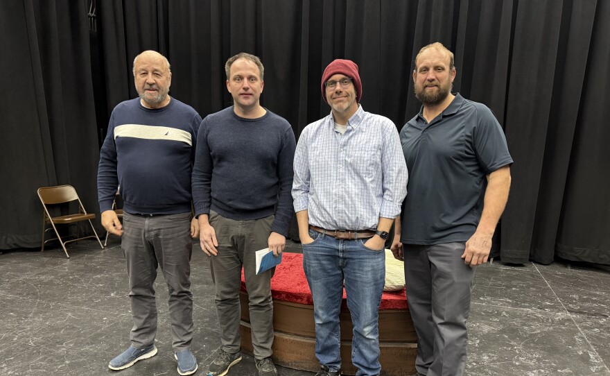 Four men standing on stage with a black curtain behind them.