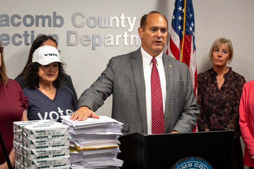 Macomb County Clerk and Republican candidate for Michigan secretary of state Anthony Forlini speaks at a podium in a county elections office. He stands before stacks of documents and boxes of copy paper, flanked by several people. American and Michigan flags stand behind him, with election-office signage on the wall.