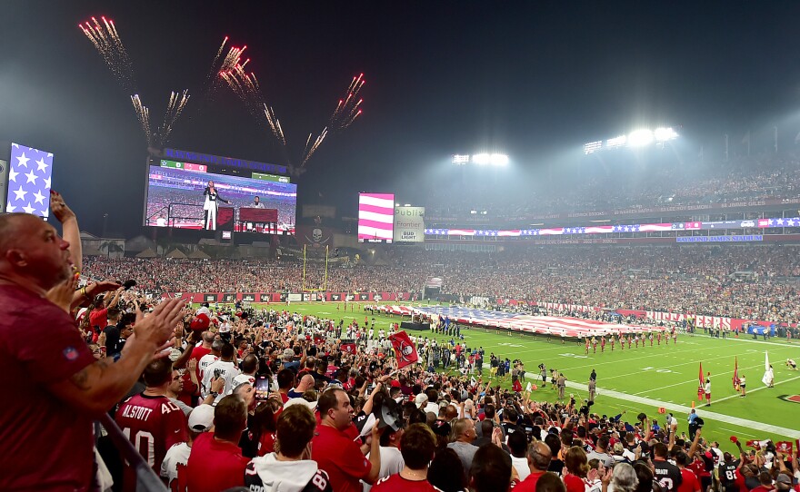 Spectators wearing red cheer in a packed football stadium, with a performance pictured on the TV screen and fireworks in the dark sky.