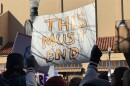 A protester holds a sign up in the air, with the words “This Must End” 