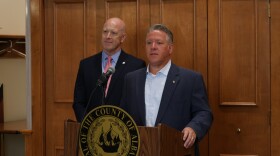 Albany County Executive Dan McCoy is speaking at a lectern with the county's seal. Albany County District Attorney Lee Kindlon is standing behind him. They're both wearing suits. The room is too boring to be worth describing. 