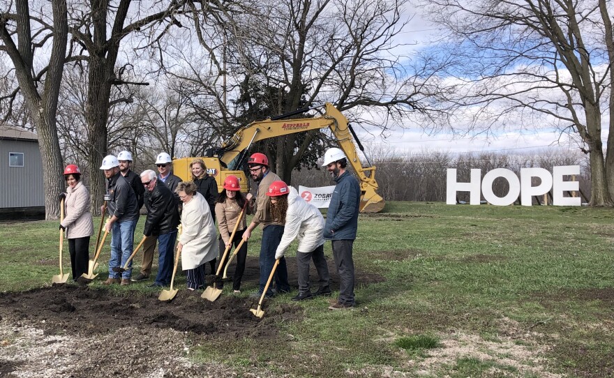 A group of men and women push gold shovels into a pile of dirt in a grass field, with trees, a construction vehicle, and large white letters spelling "hope" in the background.  