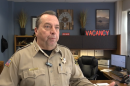 A man wearing a tan uniform and star-shaped sheriff badge speaks in his office. It's decorated with a neon sign that says "Vacancy" and some artwork of bison and a tree.