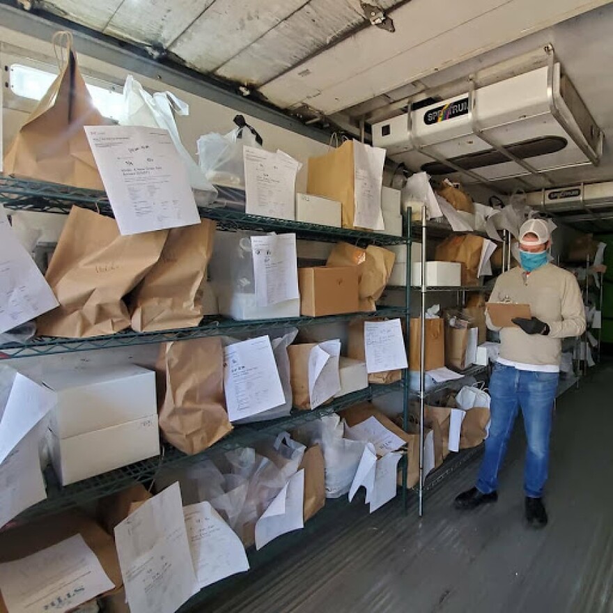 Paper to-go bags fill shelves with a person in a mask stands nearby looking at a clipboard.