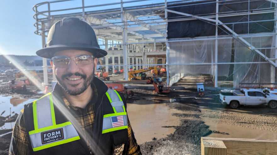A man wearing a hard had and a hi-vis vest stands in front of an under construction factory.