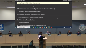A student stands alone at a podium raising his hand, facing adults on the Mat-Su school board.
